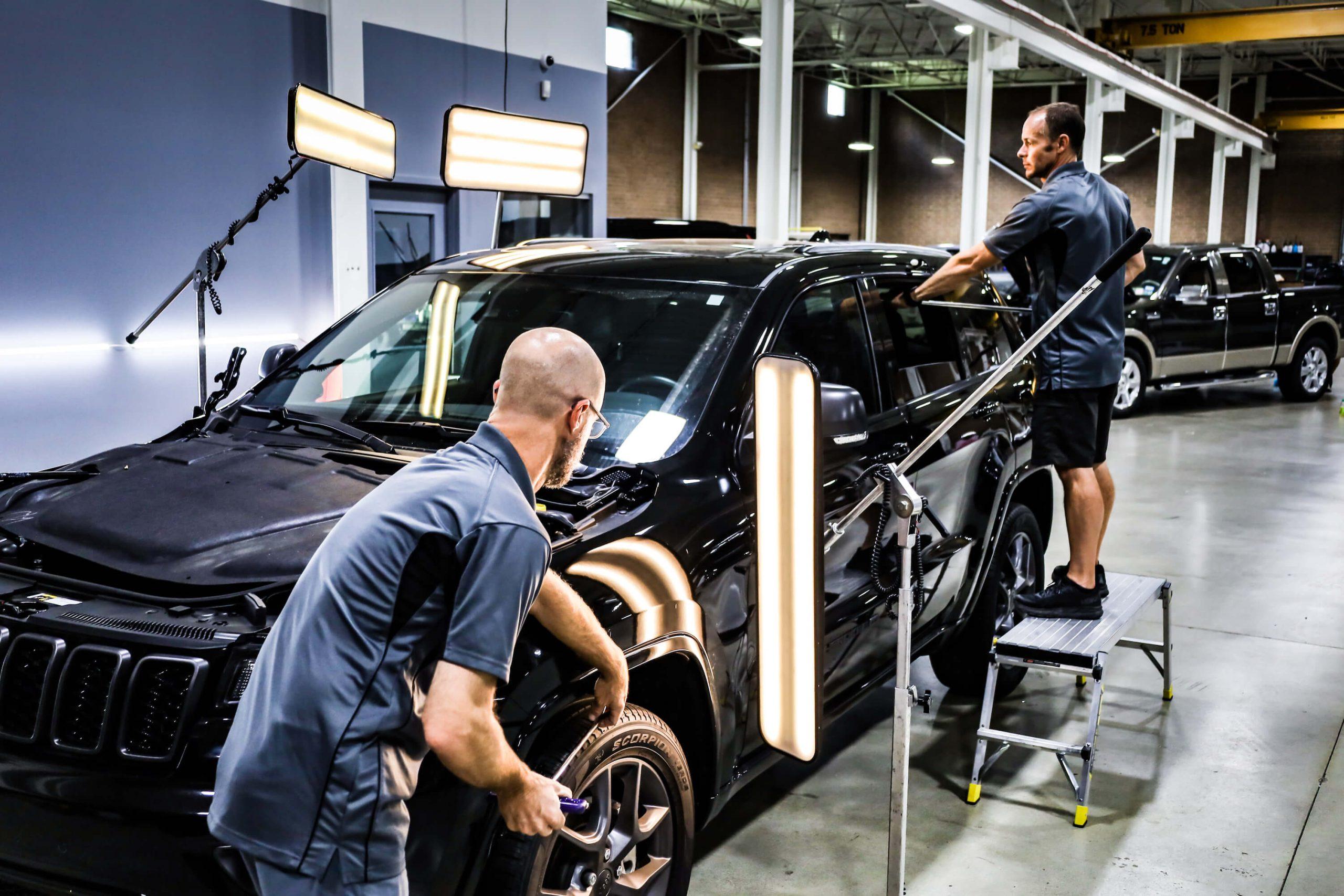 Two workers service a black SUV under bright inspection lights in an industrial automotive workshop.