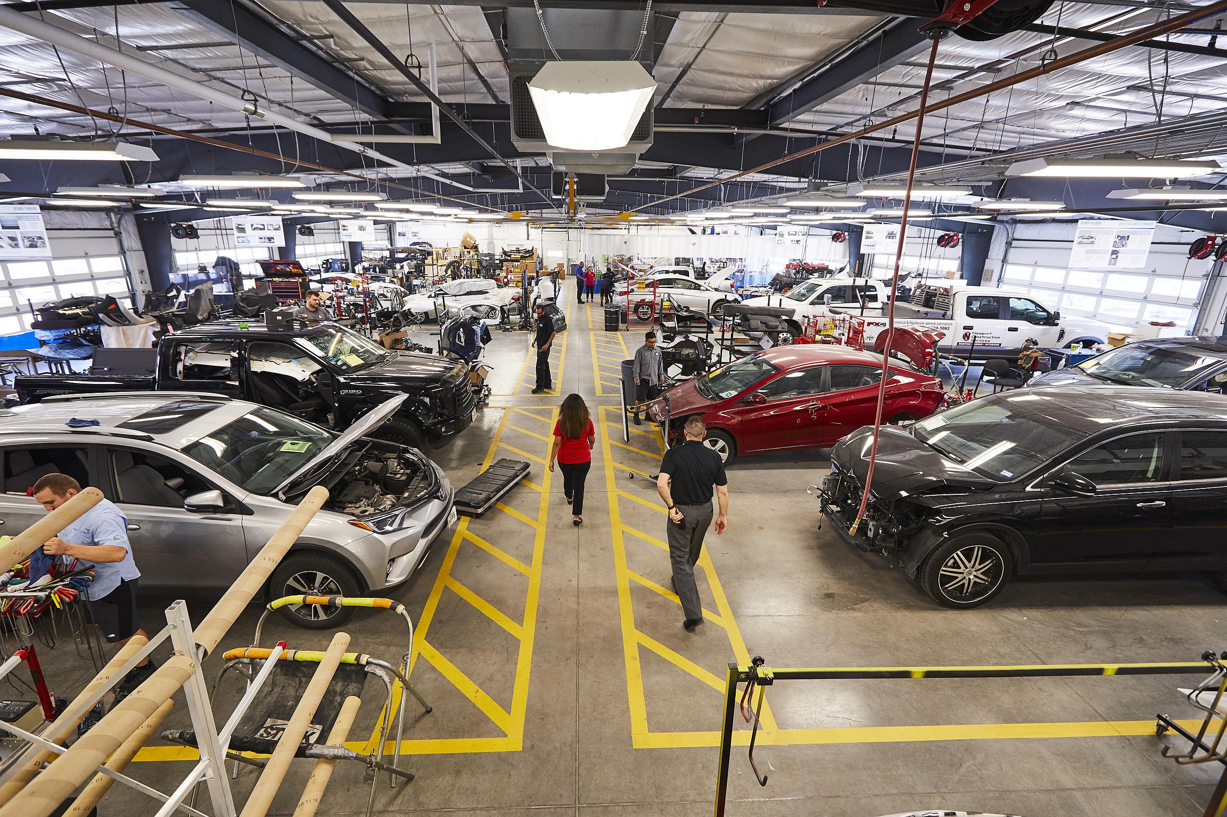 Busy automotive repair workshop filled with partially disassembled cars, workers, tools, and yellow-marked floor bays.